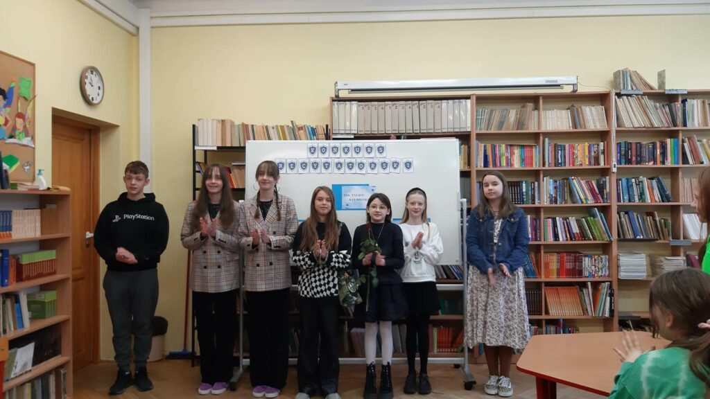 Zdjęcie zrobione w bibliotece szkolnej. W tle widać półki z książkami. Na pierwszym planie grupa uczniów za ich plecami biała tablica z napisem konkurs recytatorski. W grupie stoja dwie dziewczynku ubrane w czarne spodnie i marynarki w kratkę, które są bliźniaczkami- jedna ma rozpuszczone włosy druga dwa warkocze. Trzy dziewczynki ubrane w spódniczki lub sukienki, jedna z dzewczat w rozpuszczonych włosach ma czarne spodnie. Obok dziewcząt stoi chłopiec w czarnej bluzie z białym napisem i zielonych spodniach.