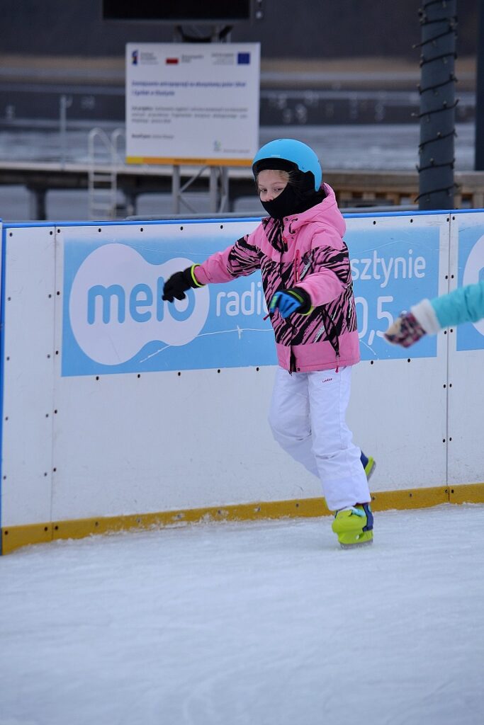 Dziewczynka w różowej kurtce jedzie na łyżwach na lodowisku. Na głowie ma niebieski kask. Ma białe spodnie i żółte łyżwy. Pod kaskiem ma czarną kominiarkę. W tle widać pomost na jeziorze.