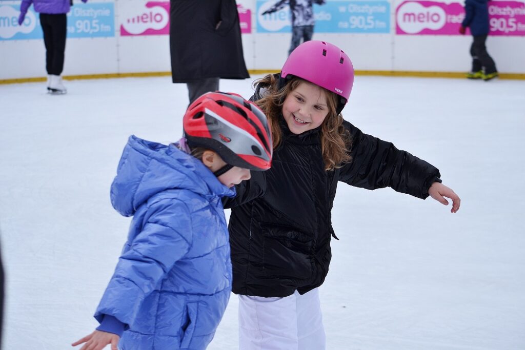 Dwie dziewczynki w kurtkach z kaskami na głowie jeżdżą na łyżwach.