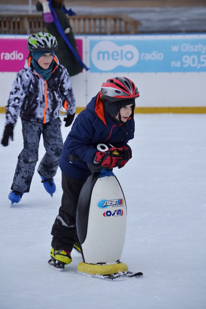 Chłopiec oparty na lodowisku o pingwinka. W tle uczennica jeżdżąca na łyżwach.
