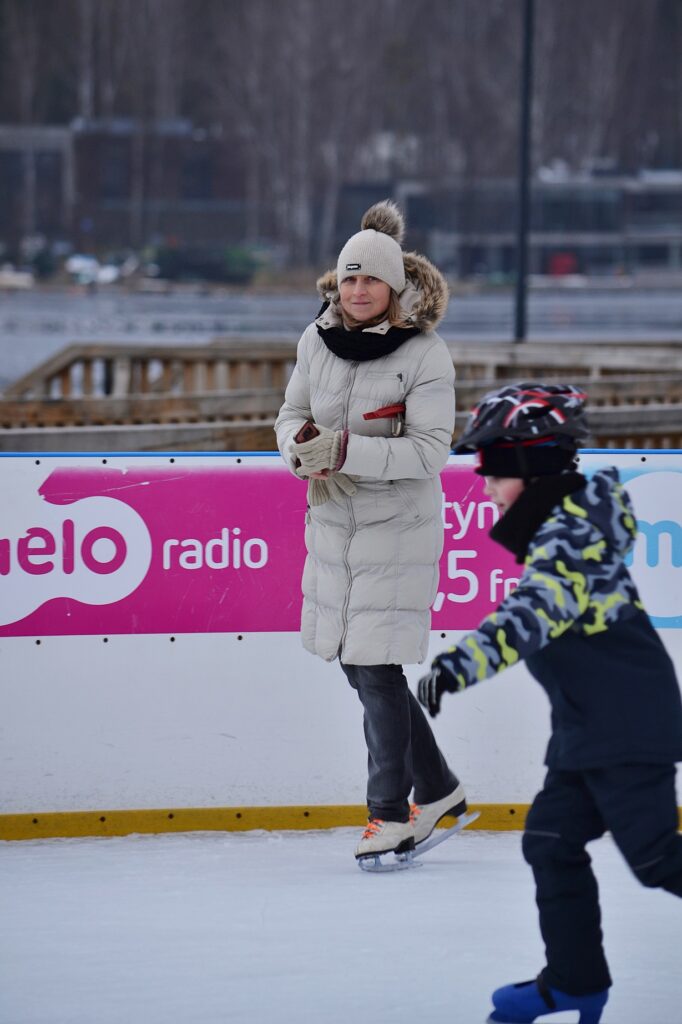 Chłopiec jeżdżący na łyżwach, ubrany w strój zimowy. W tle pani na łyżwach.