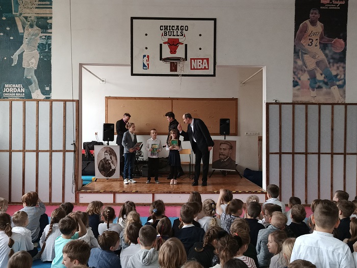 Na scenie stoi mężczyzna oraz troje uśmiechniętych dzieci, trzymających w dłoniach książki. Chłopiec z lewej strony ubrany jest w granatowe spodnie i białą koszulę. Dziewczynka w środku ma na sobie granatową spódniczkę i białą bluzeczkę. Chłopiec z prawej strony ma granatowe spodnie i białą koszulę. Mężczyzna z prawej strony ubrany jest w czarny garnitur i białą koszulę.