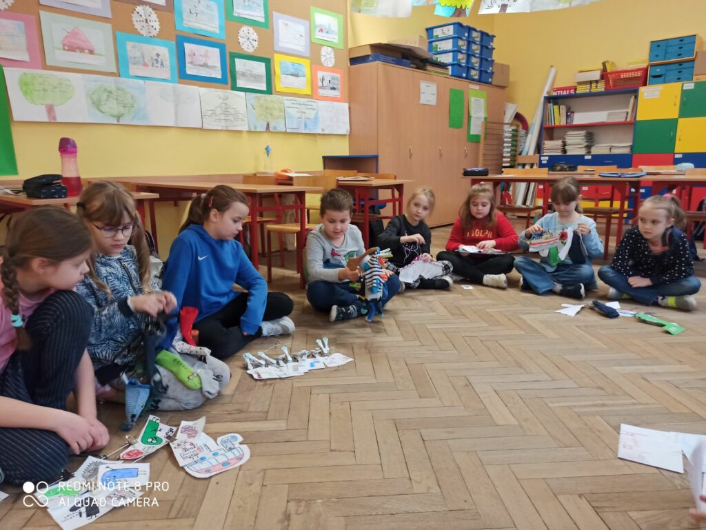 Na zdjęciu obecne są dzieci, siedzące w kole i pokazujące swoje projekty do kamery. Przedstawiają wieszaki z kolorowymi skarpetami kolegom i koleżankom z klasy. Opowiadają o nich, a po uśmiechach, wymalowanych na ich twarzach, możemy domyślić się, że są zadowoleni ze swoich prac.