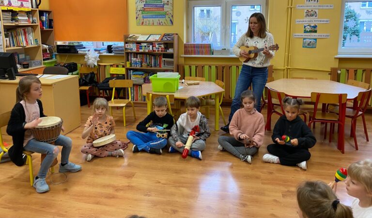 Z lewej dziewczynka w czarnym swetrze grająca na djembe. Obok dziewczynka grająca na bębenku. Chłopiec w czarnej bluzie grający na tarce, obok chłopiec w białej koszulce i szarych spodniach grający na tarce, z prawej dziewczynka w różowej bluzie grająca na żabie akustycznej, z prawej dziewczynka ubrana na czarno grająca na grzechotkę, z prawej dziewczynki, jedna w białej bluzce gra na grzechotce. W tle okna, stoliki i regał z kaiążkami. Przy oknie pani w białym swetrze i niebieskich spodniach, gra na ukulele.