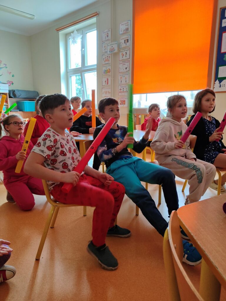 grupa dzieci siedzących na podłodze, każe z nich trzyma w ręku kolorową rurkę, która w zależności od koloru wydaje inny dźwięk.