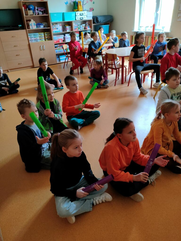 grupa dzieci siedzących na podłodze, każe z nich trzyma w ręku kolorową rurkę, która w zależności od koloru wydaje inny dźwięk.