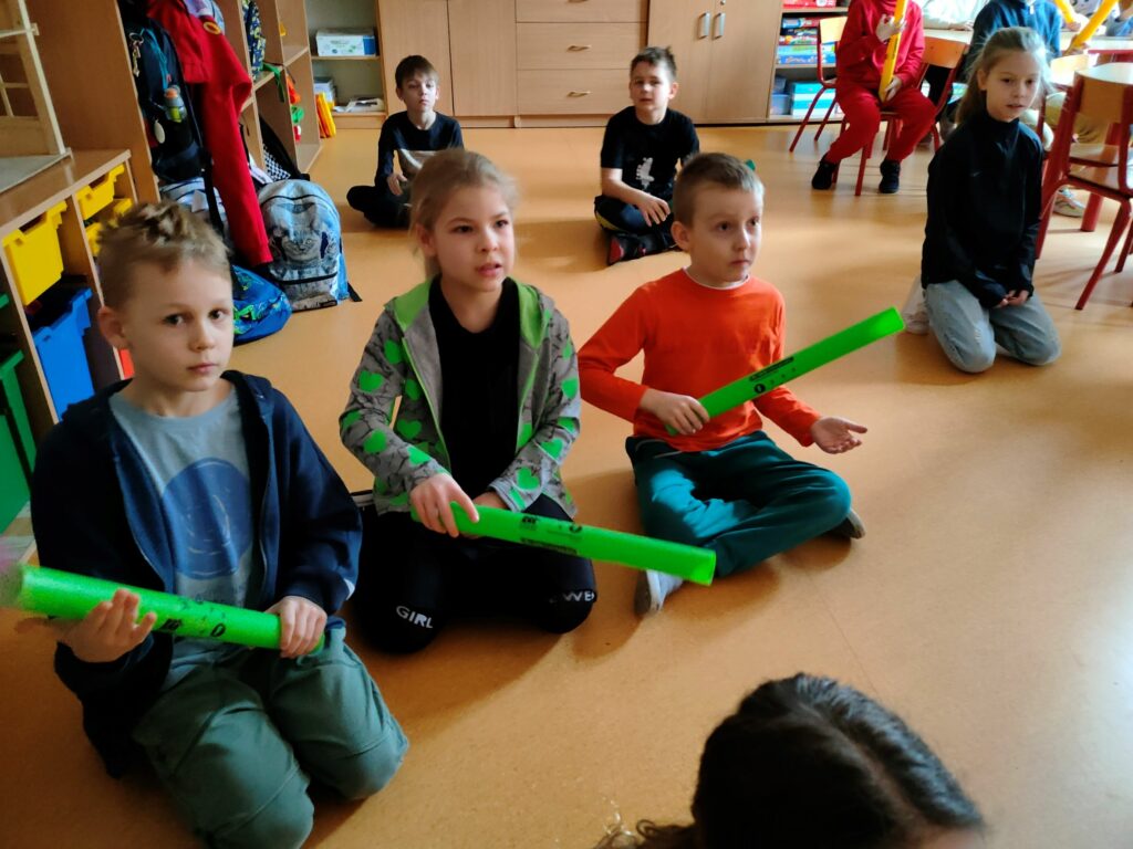 trojka dzieci siedzących na podłodze. Od lewej chłopiec w granatowej bluzie trzyma zieloną rurkę, dziewczynka w środku w zielonej bluzie trzyma zieloną rurkę, chłopiec w pomarańczowej koszulce trzyma zieloną rurkę,