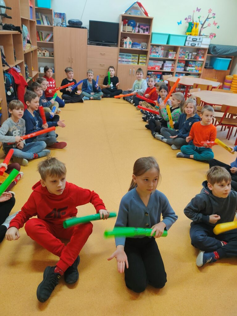 grupa dzieci siedzących na podłodze, każe z nich trzyma w ręku kolorową rurkę, która w zależności od koloru wydaje inny dźwięk.