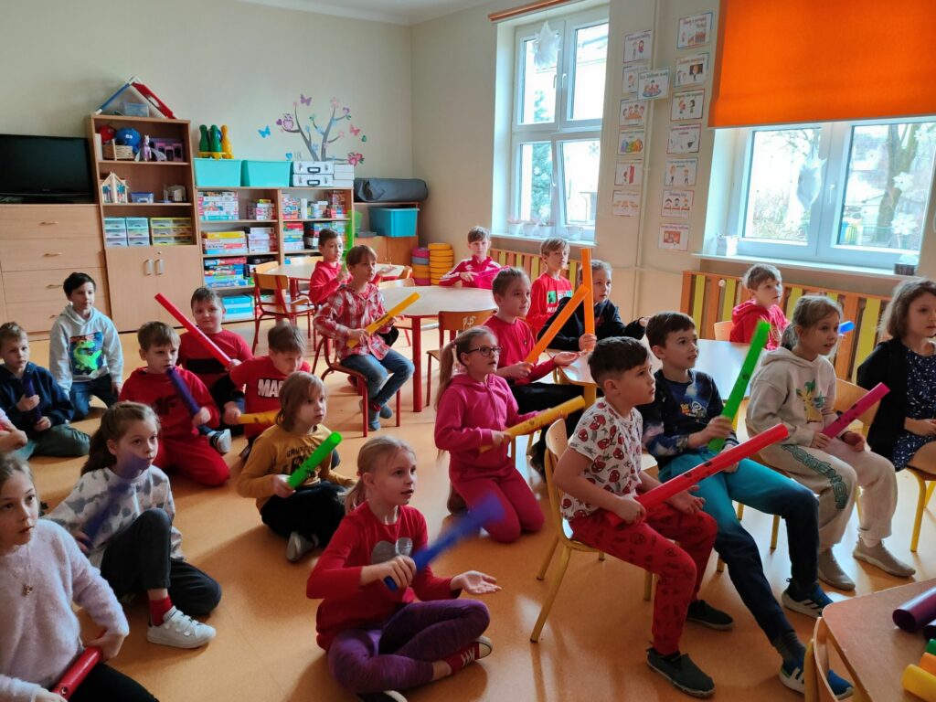 grupa dzieci siedzących na podłodze, każe z nich trzyma w ręku kolorową rurkę, która w zależności od koloru wydaje inny dźwięk.