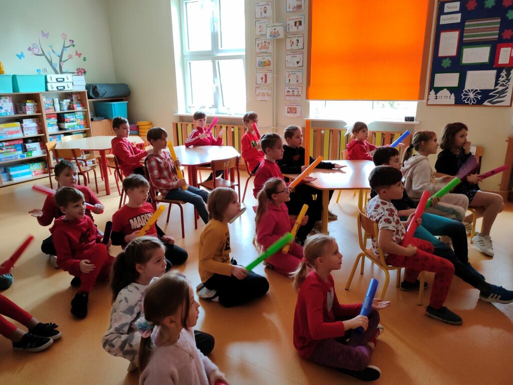 grupa dzieci siedzących na podłodze, każe z nich trzyma w ręku kolorową rurkę, która w zależności od koloru wydaje inny dźwięk.