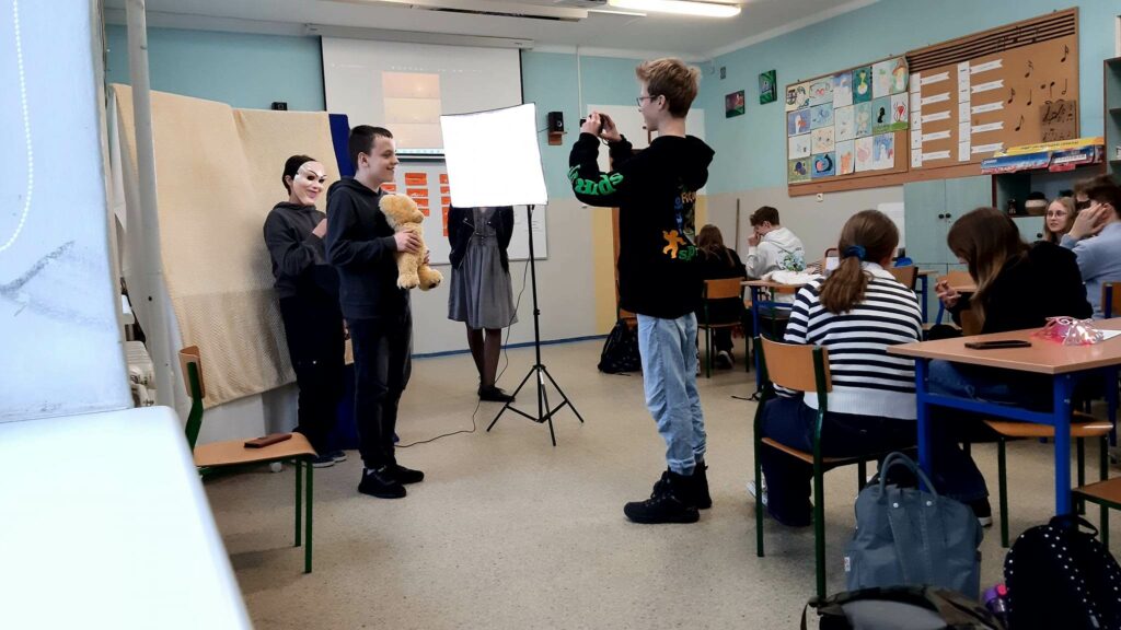 Zdjęcie przedstawia salę lekcyjną.Zdjęcie zrobione z końca sali. Po prawej stronie ławki, w których siedzą uczniowie tyłem do fotografa. Centrum zdjęcia przedstawia dwóch chłopców ustawiających się do zdjęcia oraz trzeciego robiącego zdjęcie. Chłopiec stojacy z przodu trzyma misia chłopiec stojący za nim ma na twarzy maskę mężczyzny. Na zdjęciu widać lampę oświetleniową na statywie.