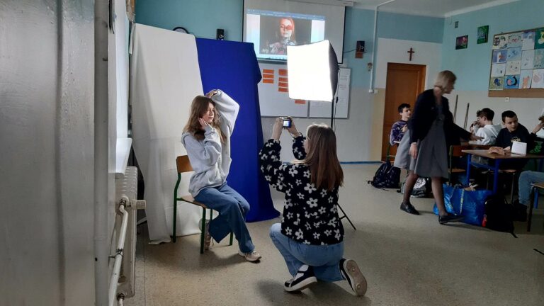 Zdjęcie przedstawia uczennice robiące zdjęcie . Jedna siedzi na krześle, rękoma obejmuje głowę, ma na sobie jasną bluzę i granatowe spodnie. Krzesło na którym siedzi ustawione jest na tle biało niebieskim. Uczennica oświetlona jest lampą fotograficzna na statywie. Druga dziewczyna klęczy na jednym kolanie jest ustawiona przodem do modelki a tyłem do fotografa. Trzyma w dłoniach aparat lekko w górze ustawiony do zrobienia zdjęcia.