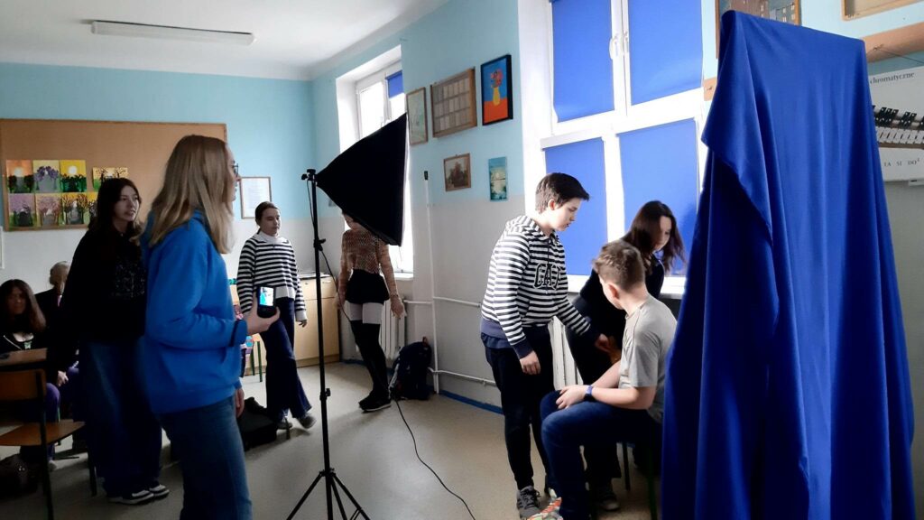 Zdjęcie przedstawia grupę uczniów przygotowujących się do zrobienia zdjecia. Jeden uczeń siedzi bokiem do fotografa, jeden stoi przodem do siedzącego, trzecia osoba to dziewczyna pochylająca się do przodu. Obok stoi lampa na statywie za lampę są dwie dziewczynki przyglądające się scenie. Obok lampy widać częściowo dziewczynę o jasnych włosach w niebieskiej bluzie trzymająca aparat fotograficzny.