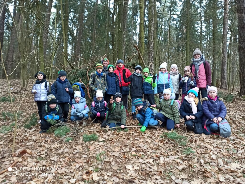 Na zdjęciu widoczni są uczniowie, którzy chwilę wcześniej mieli okazję przejść przez środek szałasu wybudowanego w środku lasu. Dzieci miały przy tym niewyobrażalną frajdę, a uśmiechy na ich twarzach mogą to tylko potwierdzić.