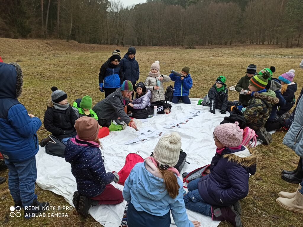 Zdjęcie pokazuje uczniów, siedzących w kole razem z panią prowadzącą wyprawę. Dzieci siedzą na białej macie. Oglądają ilustracje tropów zwierząt leśnych.