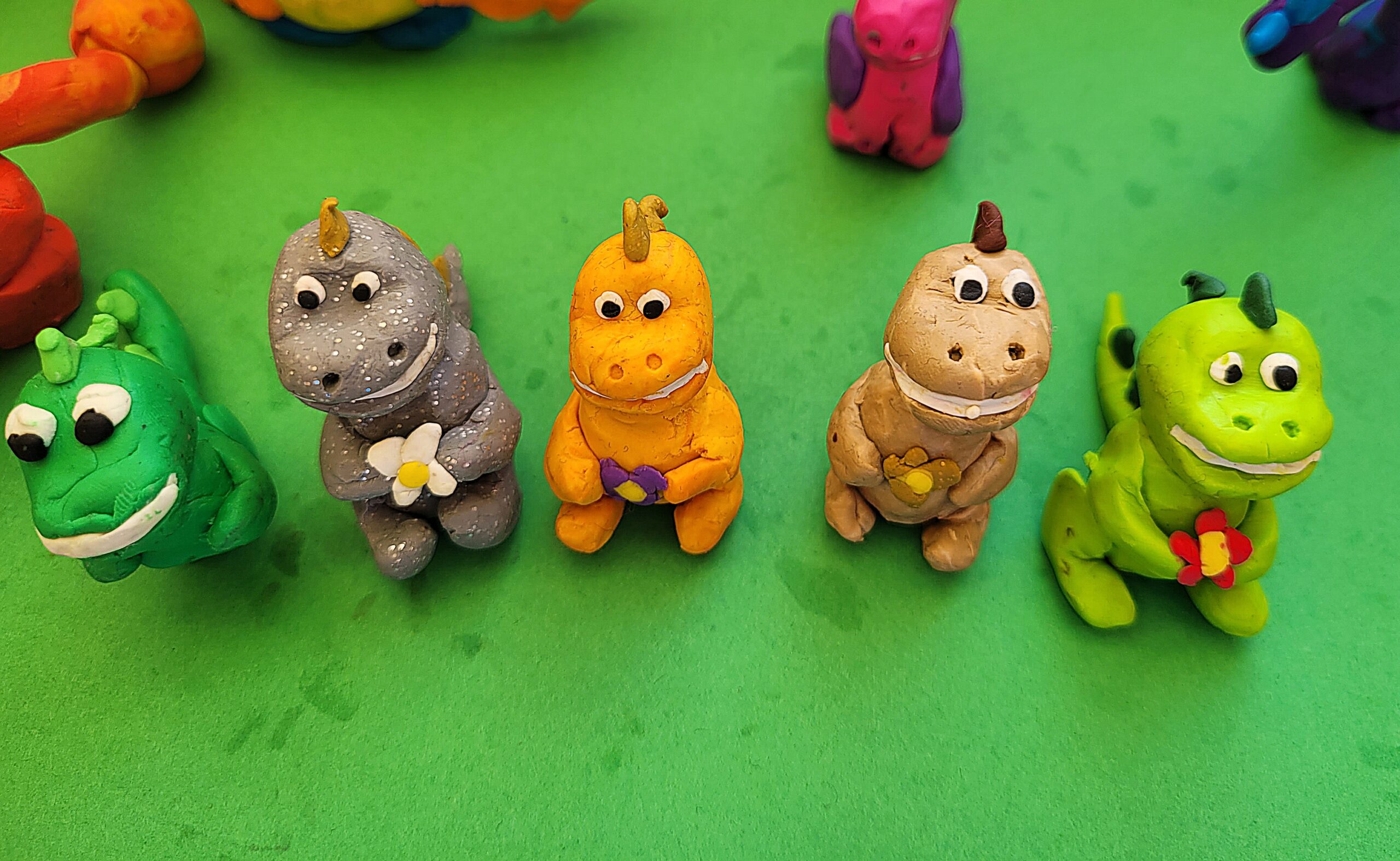 Na zdjęciu znajduje się pięć figurek dinozaurów ulepionych z plasteliny. Pierwszy dinozaur od prawej jest koloru jasnozielonego z ciemnozielonym grzbietem i trzyma w łapkach czerwony kwiatek. Drugi od prawej jest koloru złotego z brązowym grzbietem a włapkach trzyma beżowy kwiatek. Trzeci dinozaur od prawej jest koloru pomarańczowego ze złotym grzbietem i trzyma w łapkach filetowy kwiatek. Czwarty dinozaur od prawej jest koloru siwego w drobne białe kropeczki, ma złoty grzbiet i w łapkach trzyma biały kwiatek. Piąty dinozaur od prawej jest koloru zielonego i ma jasnozielony grzbiet. Wszystkie dinozaury mają szeroki biały usmiech, wyłupiaste oczy i znajdują się na zielonej kartce.