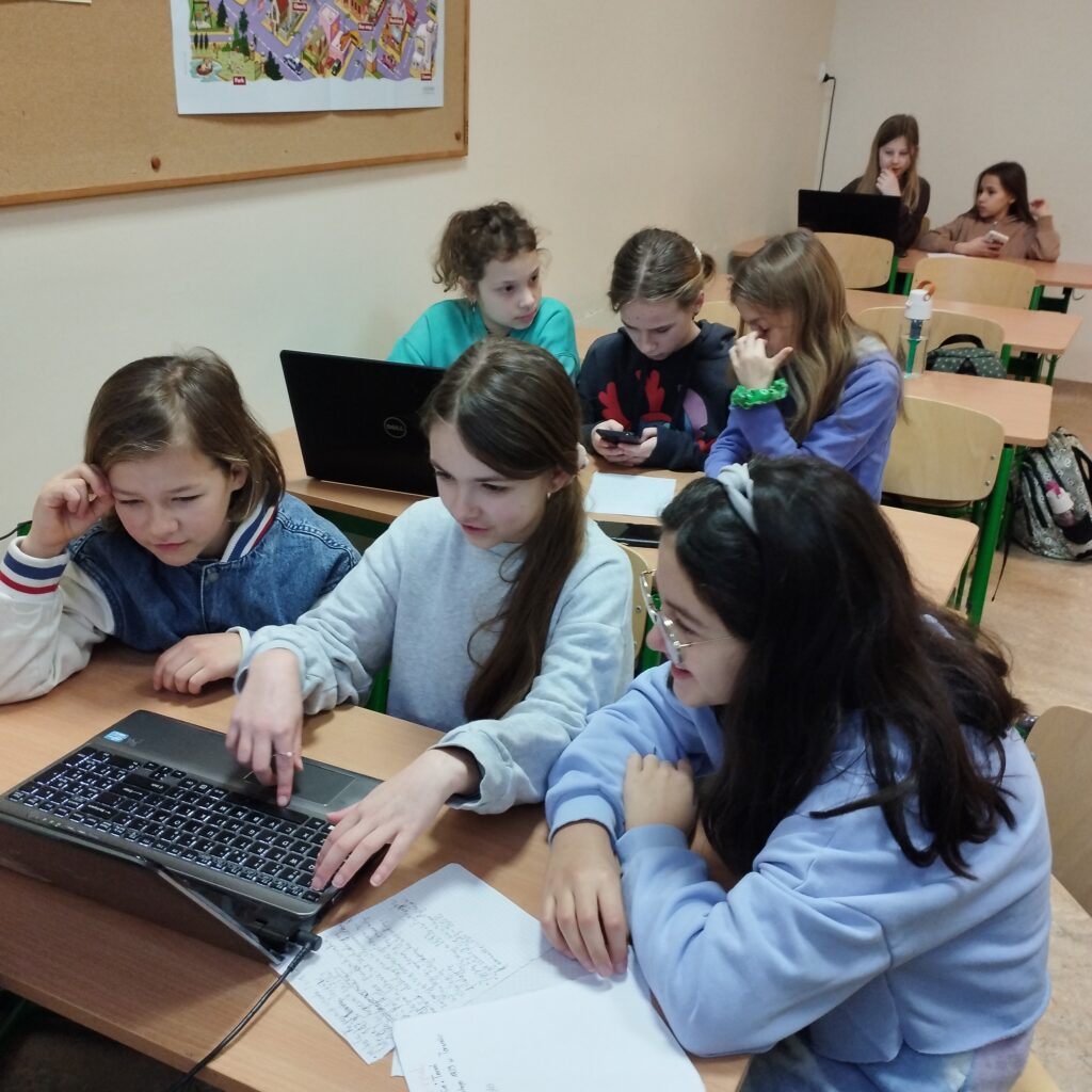 Na pierwszym planie w ławce siedzą dwie dziewczynki i chłopiec, wszyscy pracują przy laptopie, za nimi w ławce siedzą trzy dziewczynki również pracujące na laptopie, jedna z nich trzyma telefon w dłoni. Z tyłu klasy widać jeszcze dwie dziewczynki siedzące razem w ławce, przed laptopem