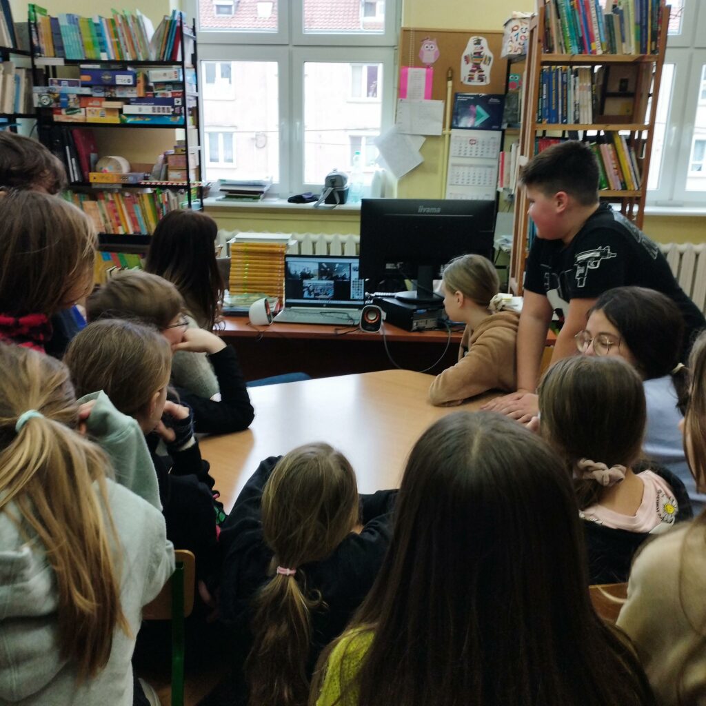 grupa dzieci siedzi w bibliotece, dookoła komuptera i patrzy w monitor