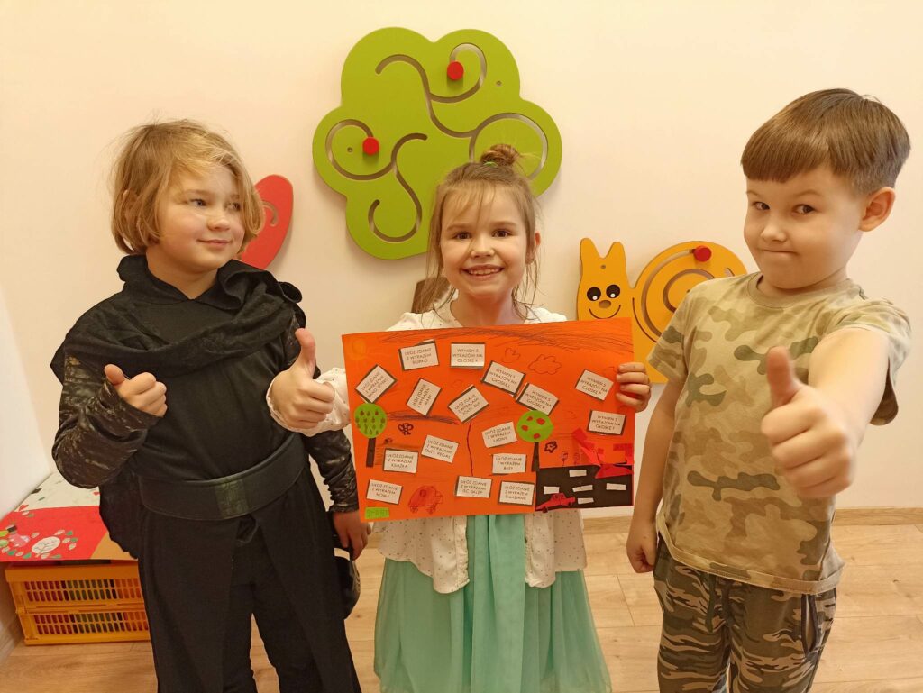 Po lewej stronie stoi chłopiec, który pokazuje kciuk w górę. Lekko spogląda na stojącą obok niego dziewczynkę, która ma uniesiony prawy kciuk w górę, a w lewej ręce trzyma pomarańczową kartkę A3, która prezentuje grę logopedyczną z zadaniami. Obok dziewczynki po prawej stronie stoi chłopiec z uniesionym kciukiem w górę i dumnym wyrazem twarzy.