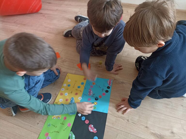 Na pierwszym planie znajduje się kolorowa plansza do gry z zadaniami, która leży na podłodze. Nad nią znajdują się trzej chłopcy. Chłopiec po lewej stronie przestawia pionek na planszy. Chłopiec znajdujący się na środku, rzuca kostką do gry. Trzeci chłopiec po prawej stronie obserwuje grę kolegów.