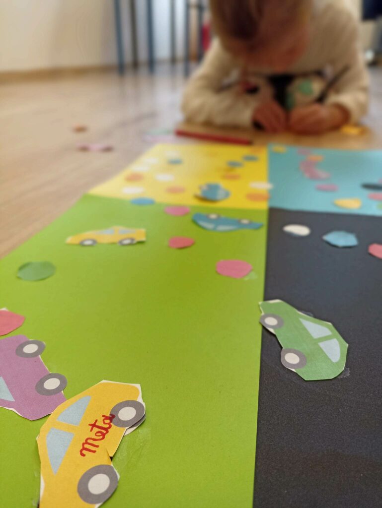 Na pierwszym planie znajduje się plansza do gry, która składa się czterech połączonych kartek a4 w kolorze zielonym, niebieskim, żółtym i czarnym. Na planszy znajdują się wycięte kolorowe kółka oraz kolorowe samochodziki. Na żółtym samochodziku widnieje napis „meta”. W tle widać konury pochylonej dziewczynki, która pisze zasady gry na kartach.