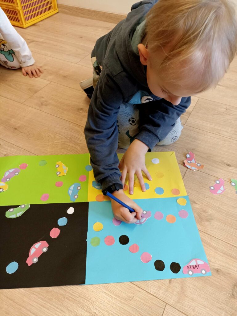 Zdjęcie przedstawia chłopca o blond włosach, który jest pochylony nad planszą do gry. W prawej ręce trzyma niebieski flamaster. Chłopiec na fioletowym samochodziku zapisuje znak plus. Plansza do gry leży na podłodze przed chłopcem. Składa się czterech połączonych kartek a4 w kolorze zielonym, niebieskim, żółtym i czarnym. Na planszy znajdują się wycięte kolorowe kółka oraz kolorowe samochodziki.
