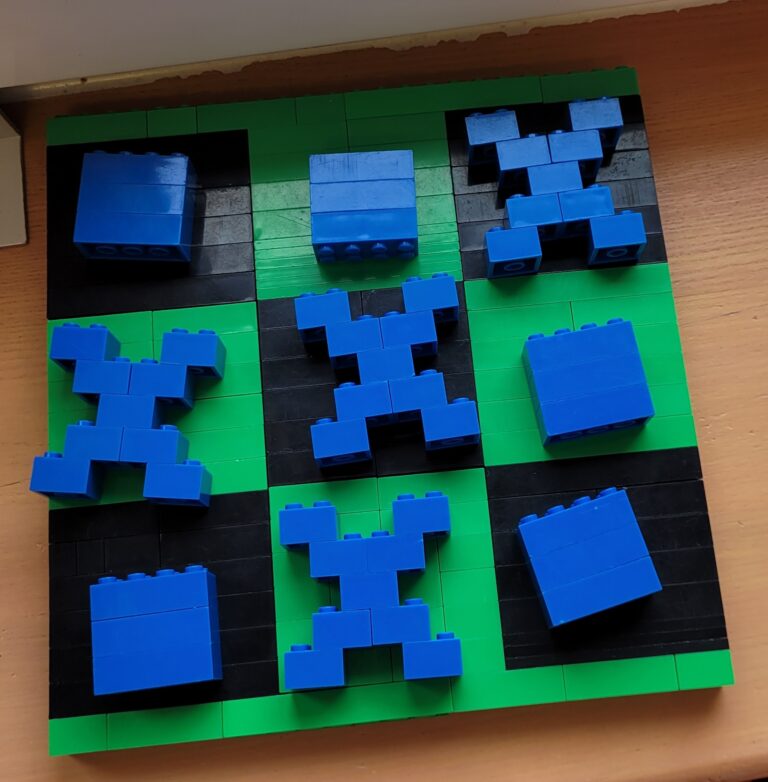 Na zdjęciu znajduje się kwadratowa zielono- czarna plansza imitująca planszę do gry w kółko i krzyżyk. Na planszy znajdują się figurki imitujące kółko i krzyżyk. Plansza i pozostałe elementy zbudowane są z klocków Lego.