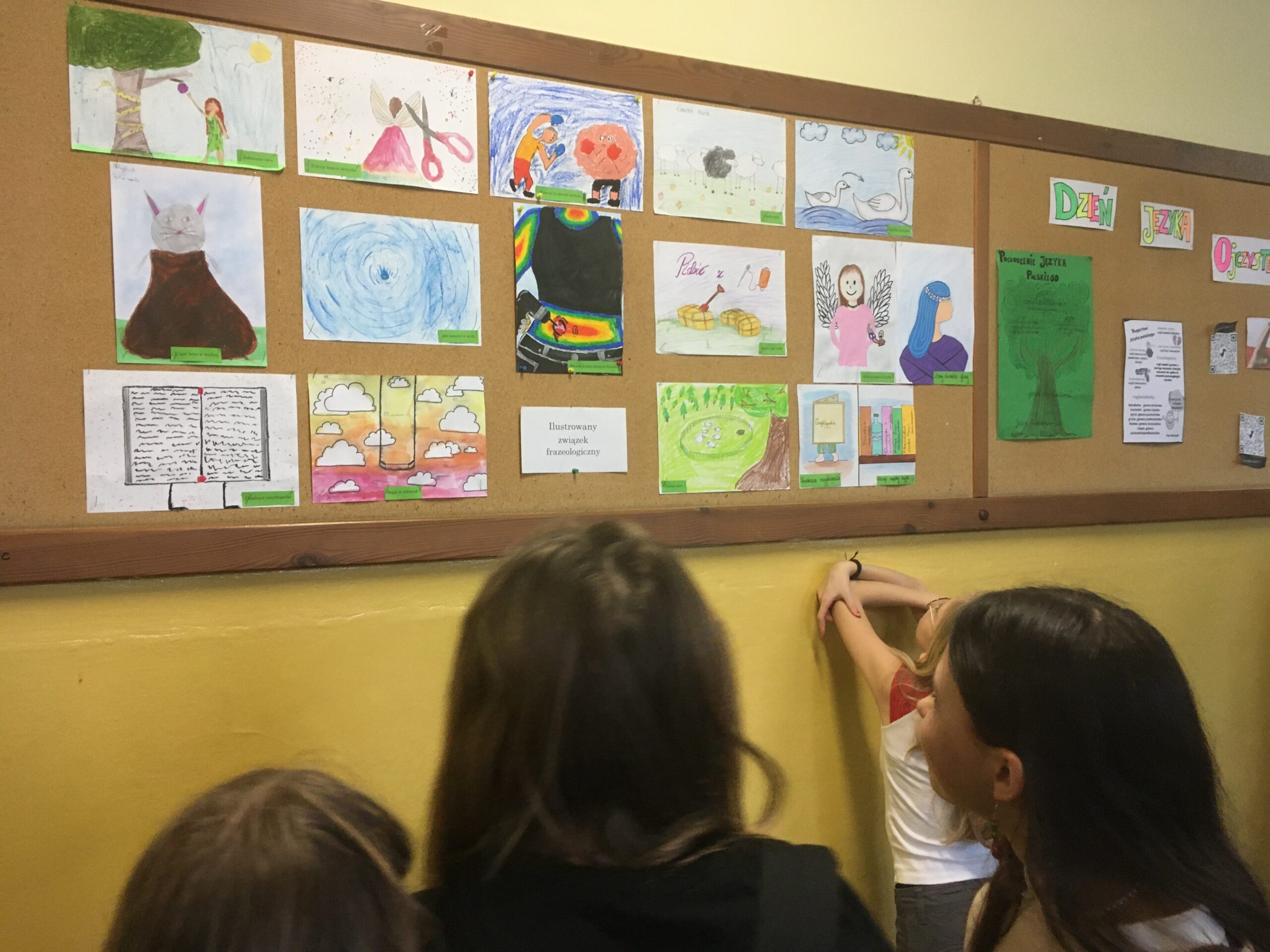 Zdjęcie przedstawia szkolny korytarz. Cztery uczennice odwrócone tyłem i patrzą na gazetkę ścienną. Na gazetce z lewej strony znajduje się kilkanaście prac plastycznych ilustrujących związki frazeologiczne. Na dole po środku jest podpis: „Ilustrowany związek frazeologiczny”. Z prawej strony widać oddzielony drewnianą ramą fragment napisu „Dzień Języka Ojczystego” oraz zielony plakat przedstawiający drzewo języków słowiańskich i kartkę zawierającą ciekawostki językowe, obok znajdują się kody QR.