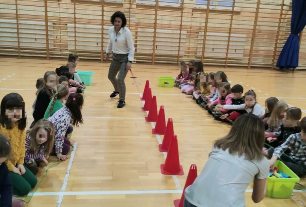 Na zdjęciu widać 2 rzędy dzieci oddzielonych od siebie rzędem czerwonych pachołków. Dzieci siedzą na podłodze. Na każdym szczycie rzędów widać po jednej kobiecie. Jedna kuca, druga stoi w lekkim rozkroku