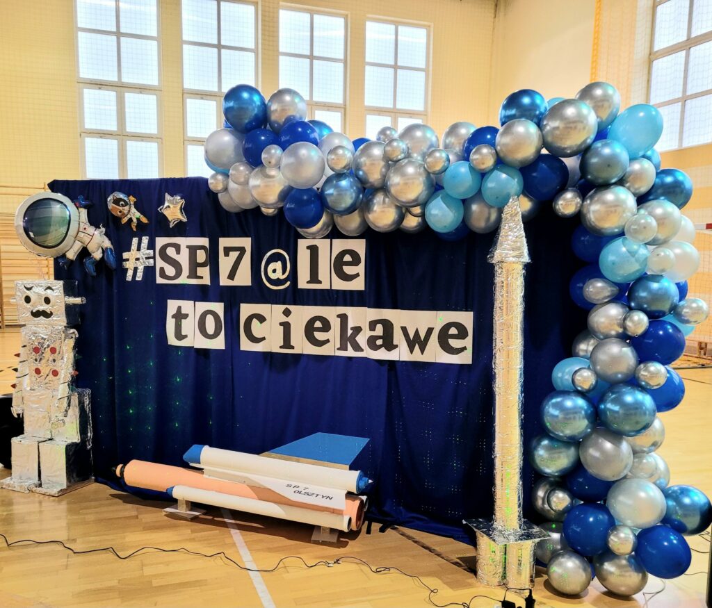 Na zdjęciu widoczna jest ścianka ze scenografią- wizytówką Dnia Otwartego Szkoły SP7. Od połowy, wzdłuż górnej i bocznej ramy ciągnie się girlanda z finezyjnie połączonych balonów różnej wielkości i kolorystyce (perłowe, niebieskie i granatowe połyskujące). W środkowej części ścianki na granatowym tle przyczepiony jest napis #SP7@le to ciekawe. Pod nim, na podłodze położony jest wahadłowiec kosmiczny w kolorach białym, niebieskim i kremowym. Po jego prawej stronie na podłodze stoi tuba na kształt rakiety w srebrnej folii aluminiowej. Po lewej zaś stronie stoi robot z kartonów, pokryty folią aluminiową z twarzą wąsacza, odstającymi uszami z rolek i długimi, spiralnymi rękoma. Nad nim, w górnym rogu ścianki przyklejone są trzy zabawne, kosmiczne stworki.