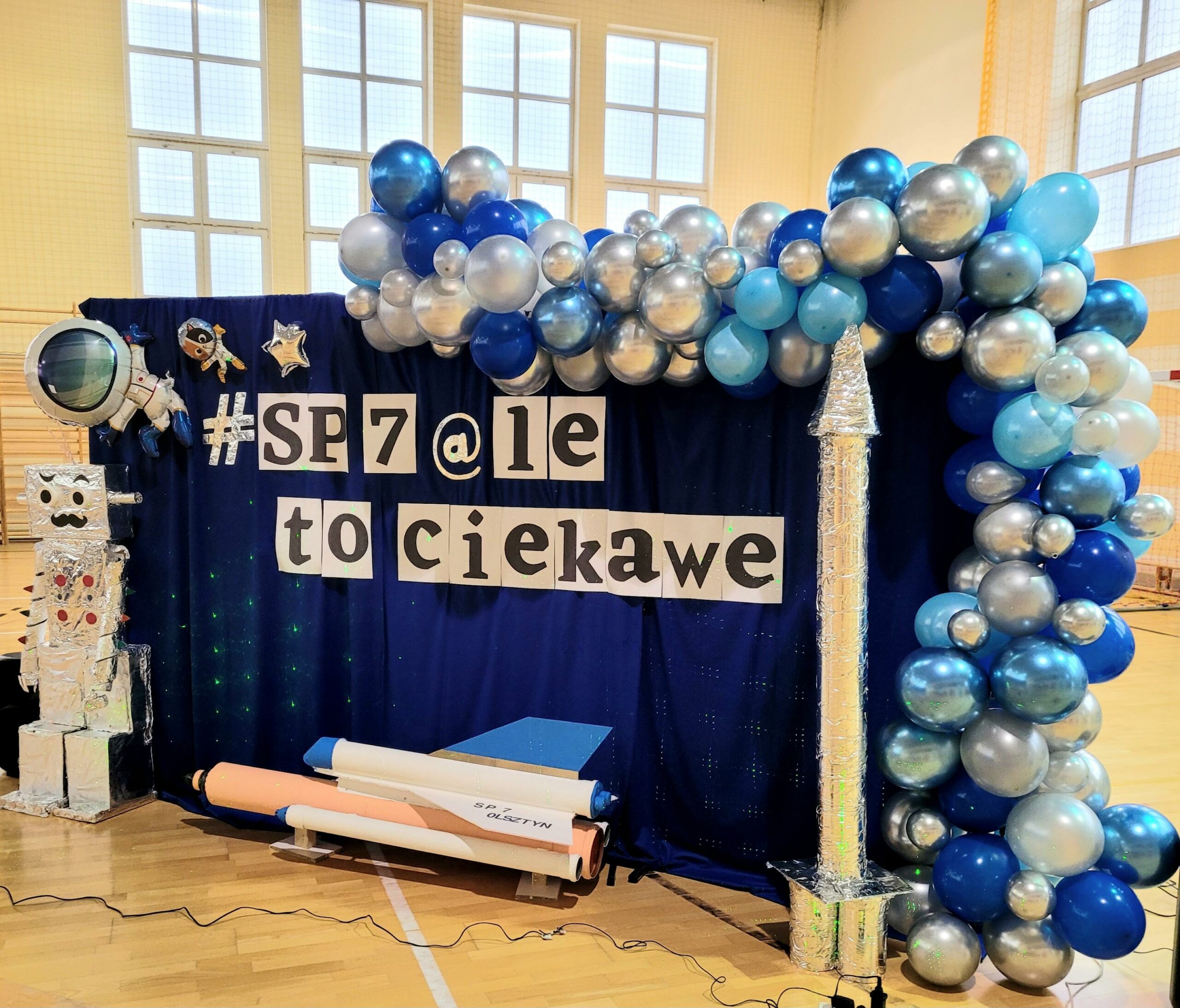 Na zdjęciu widoczna jest ścianka ze scenografią- wizytówką Dnia Otwartego Szkoły SP7. Od połowy, wzdłuż górnej i bocznej ramy ciągnie się girlanda z finezyjnie połączonych balonów różnej wielkości i kolorystyce (perłowe, niebieskie i granatowe połyskujące). W środkowej części ścianki na granatowym tle przyczepiony jest napis #SP7@le to ciekawe. Pod nim, na podłodze położony jest wahadłowiec kosmiczny w kolorach białym, niebieskim i kremowym. Po jego prawej stronie na podłodze stoi tuba na kształt rakiety w srebrnej folii aluminiowej. Po lewej zaś stronie stoi robot z kartonów, pokryty folią aluminiową z twarzą wąsacza, odstającymi uszami z rolek i długimi, spiralnymi rękoma. Nad nim, w górnym rogu ścianki przyklejone są trzy zabawne, kosmiczne stworki.