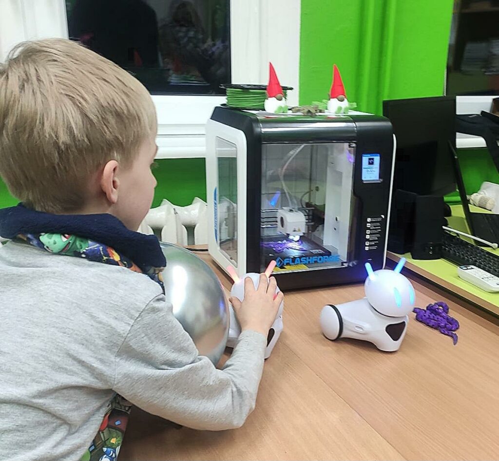 Na pierwszym planie jest chłopiec oparty łokciami o ławkę. Prawą ręką dotyka niewielki robot w kształcie zwierzątka z czółkami. Chłopiec z zainteresowaniem obserwuje pracę drukarki 3D.