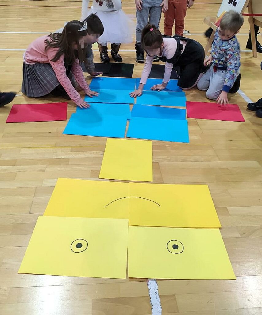 Zdjęcie przedstawia czworo dzieci podczas aktywności - klęczą lub są pochylone i układają postaci z kolorowych kartek A3. Głowa składa się z 4 żółtych kartek z narysowanymi oczami i szerokim uśmiechem, szyja to jedna kartka żółta, tułów z 8 kartek niebieskich, ręce - po jednej czerwonej z obu stron, każda noga to jedna kartka czarna.