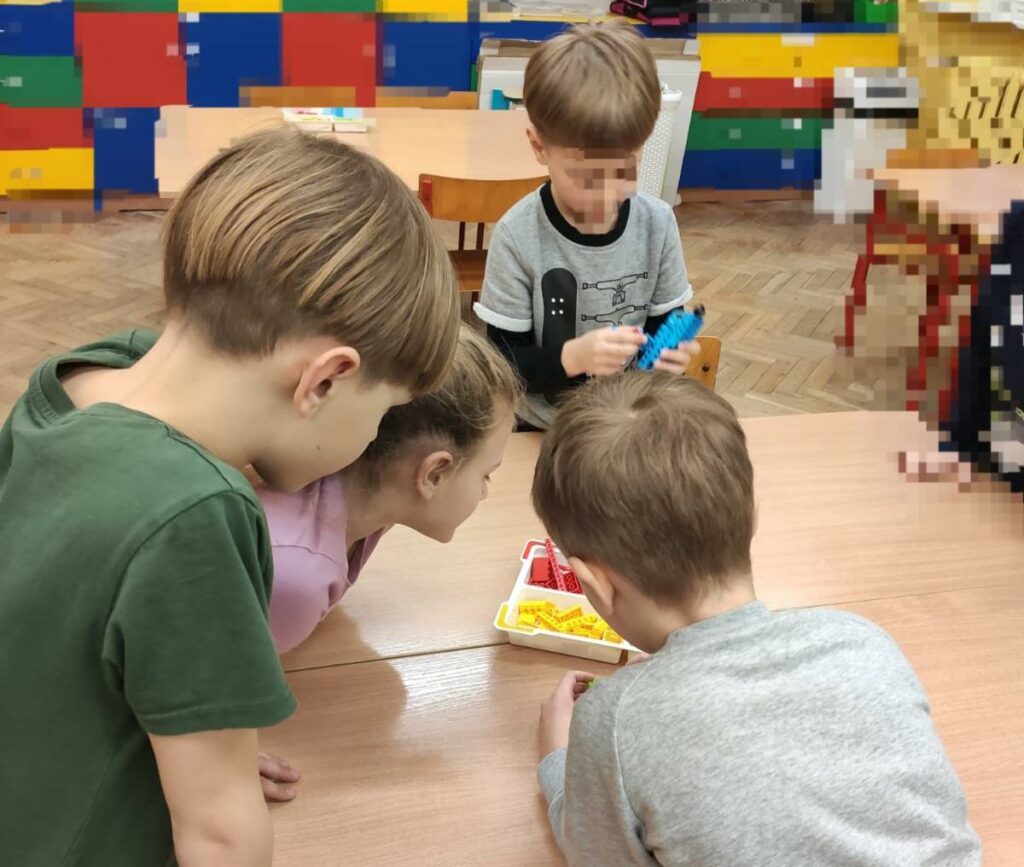 Na zdjęciu znajduję się czwórka dzieci, trzech chłopców i jedna dziewczynka. Chłopiec w szarej koszulce, zwrócony przodem do obiektywu aparatu konstruuje ludzika z niebieskich klocków lego. Dziewczynka po jego prawej stronie przygląda się co buduje. Obok dziewczynki stoi jeszcze dwóch chłopców, którzy próbują zbudować coś z czerwonych i żółtych klocków lego leżących na stoliku, wokół którego wszyscy stoją.