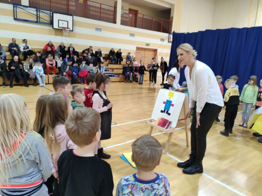 Zdjęcie zostało wykonane w Sali gimnastycznej, na trybunach widać ludzi, którzy przeszli obejrzeć Dzień Otwarty Szkoły. Na parkiecie sali gimnastycznej stoi nauczycielka, ubrana w czarno biały strój, objaśniająca dzieciom zasady zabawy. Obok niej ustawiona jest sztaluga, na której widać ludzika wykonanego z kolorowych kwadratowych kawałków papieru. Przed nauczycielką stoją zasłuchane dzieci, ubrane w kolorowe ubrania.