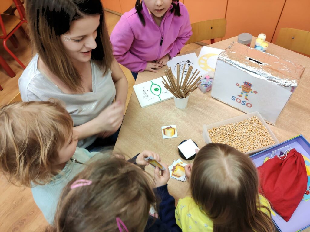 Na zdjęciu widać nauczycielkę z czwórką dzieci, są to dziewczynki, siedzą przy stoliku, na którym znajdują się słomki, obrazki, groch w pojemniku, karton z otworem, woreczek oraz bańki. Dzieci przyglądają się przedmiotom leżącym na stoliku. Jedna z dziewczynek trzyma obrazek przedstawiający prezent.