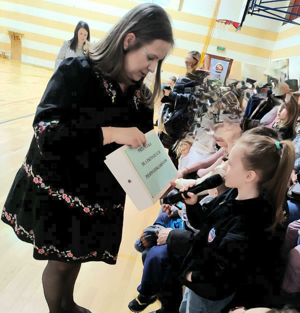 Na zdjęciu widać kobietę, ubraną w czarną sukienkę, która w ręku trzyma pudełko z naklejoną kartką z napisem "przesyłka dla przyszłych pierwszoklasistów". Pudełko jest skierowane w stronę dziewczynki, która czyta napis. W tle widać dużą grupę ludzi.