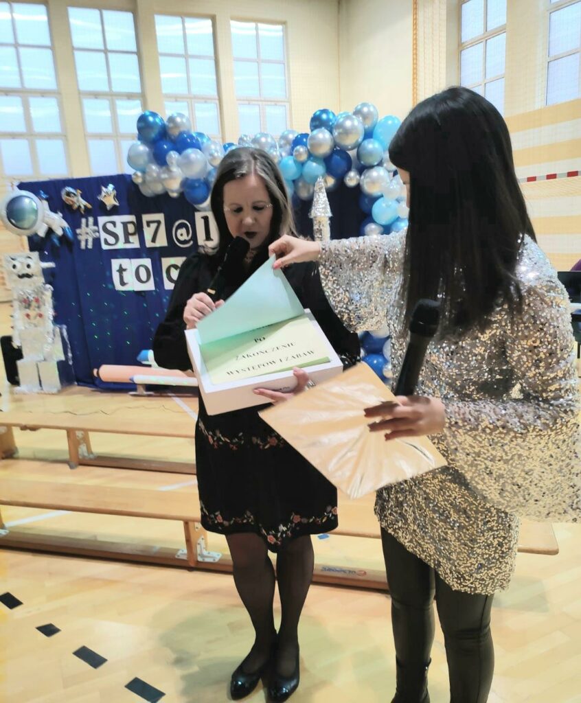 Na zdjęciu widać dwie kobiety. Kobieta po lewej stronie ubrana jest w czarną sukienkę, kobieta po prawej w srebrną i mieniącą się. Kobieta po lewej trzyma pudełko, na którym naklejone są kartki z napisami. Kobieta po prawej odsłania pierwszą kartkę i widać napis: "otworzyć po skończonej zabawie". W tle widać niebieską tablicę dekoracyjną ozdobioną girlandą balonową w kolorze niebiesko- srebrnym oraz napisem #SP @le to ciekawe.