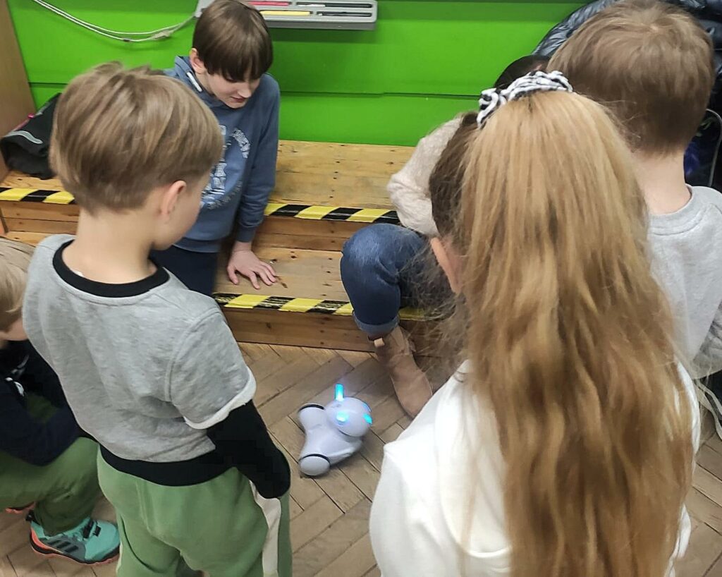 Na zdjęciu widać troje dzieci i kobietę ustawionych w kręgu. W środku znajduje się robót, który jest sterowany przez kobietę.