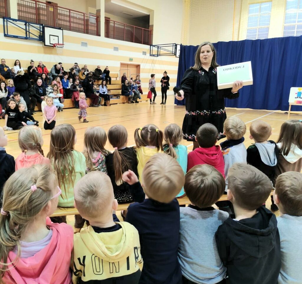 Zdjęcie przedstawia dyrektor szkoły w skierowanym w stronę uczniów mikrofonem, w drugiej ręce trzyma kartkę z napisem odkoduj. Przed nią na ławeczkach siedzą dzieci. W tle, na trybunach siedzą rodzice.