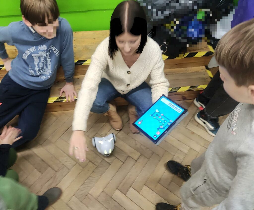 Nauczycielka w otoczeniu dzieci, w lewej dłoni trzyma tablet z włączoną aplikacją do sterowania robotem. Biały robot znajduje się na podłodze.
