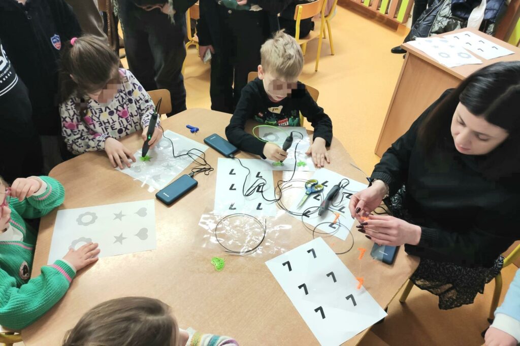 Dzieci wraz z nauczycielką zajmują miejsca dookoła stołu. Na stoliku leżą szablony, z których korzystają dzieci tworząc modele za pomocą długopisów 3d.