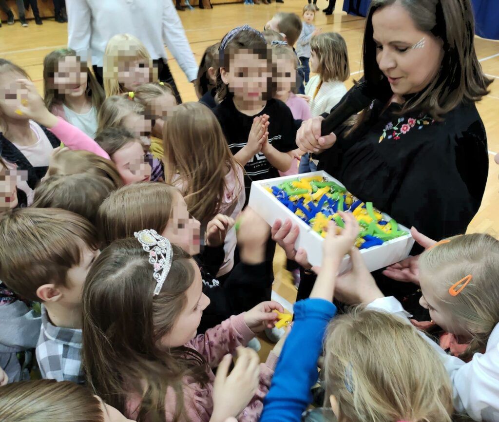 Na zdjęciu znajduje się kobieta, która trzyma w ręku białe pudełko wypełnione zielonymi, żółtymi i niebieskimi breloczkami "7". Wokół kobiety stoi duża grupa dzieci, która sięga do pudełka po breloczek.