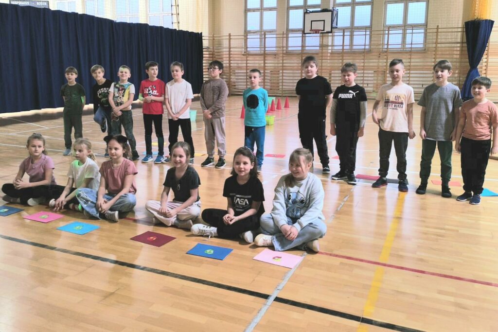 Zdjęcie wykonane jest w Sali gimnastycznej. Widzimy na nim uczniów klasy II b – dwunastu chłopców ubranych w kolorowe koszulki stoi na parkiecie w rzędzie, przed nimi siedzi sześć dziewczynek, ubranych w kolorowe koszulki. Przed każdą z dziewczynek leży kolorowa kartka.