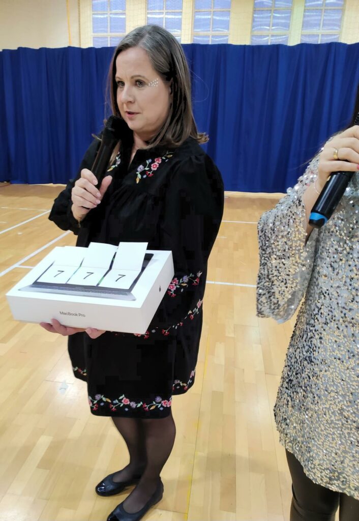 Na zdjęciu znajduje się kobieta ubrana w czarną sukienkę . W prawej ręce trzyma mikrofon, a w lewej pudełko z cyframi 777. Za kobietą widać niebieski materiał.