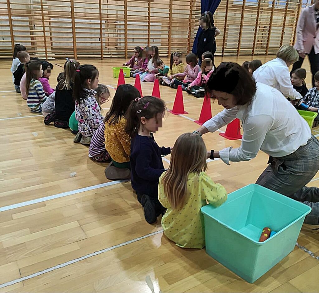 Na zdjęciu znajduje się gromada dzieci , które siedzą na podłodze na sali gimnastycznej. Obok nich widoczna jest kobieta ubrana w białą koszulę i szare spodnie. Wśród dzieci porozstawiane są czerwone pachołki oraz duże , seledynowe pudełko.