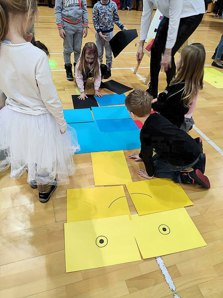 Na zdjęciu widać 6 dzieci i jedna kobietę. Dzieci ustawiono w kręgu (część z nich stoi, jedno kuca, jedno siedzi). Układają one jakąś figurę lub postać z kolorowych kartek.