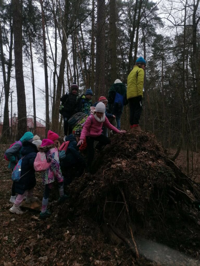 Dzieci znajdują się w lesie. Stoją na dużym korzeniu, wyrwanym z ziemi.