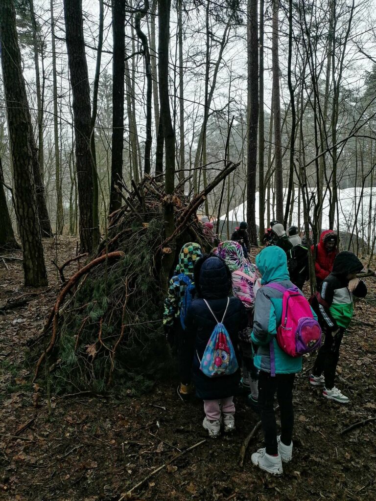 Dzieci ubrane w kurtki, stoją przed szałasem, wykonanym ze świerkowych gałęzi.