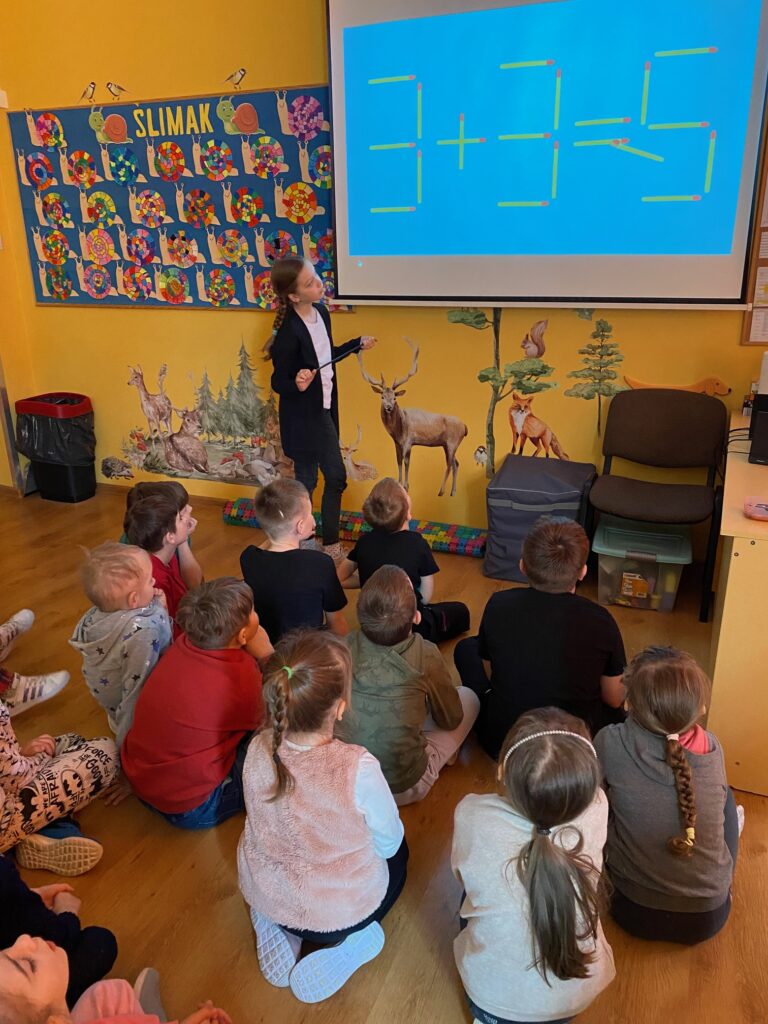 Dzieci siedzące na podłodze wpatrzone w ekran rzutnika na którym widoczne jest działanie matematyczne, ułożone z zapałek. Obok ekranu stoi dziewczynka ze wskaźnikiem w ręku.